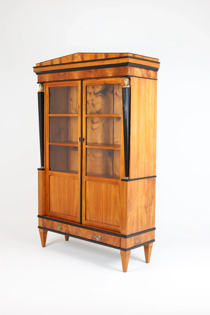 A Biedermeier vitrine from South Germany dating to 1820 to 1825 captures the regional character at its most refined. Conical feet, two doors each with four fields, three of them glazed, cherry wood veneer with very nice symmetrical grain, two ebonized pilasters with caryatids at the top and feet at the bottom, and a Schinkel-style pediment topping the case.